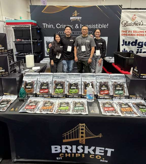 Three people standing behind a display of Brisket Chips Co. products at an event.